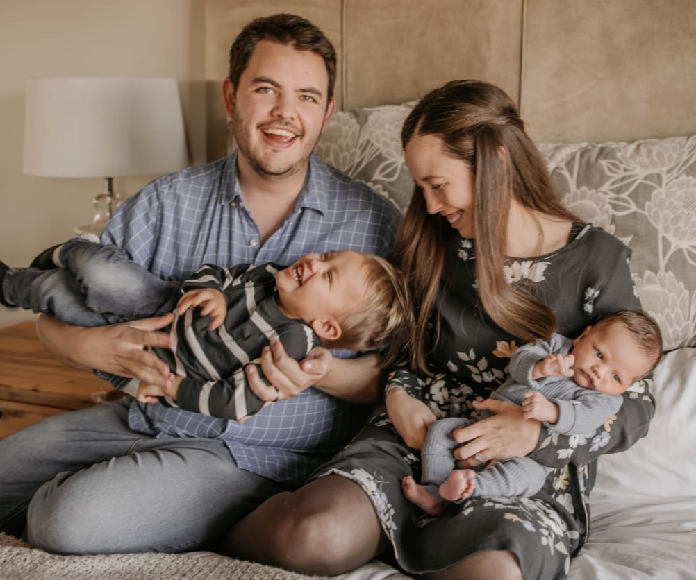 Smiling parents sitting on a bed, the father holding a laughing toddler and the mother holding a baby in a gray outfit.