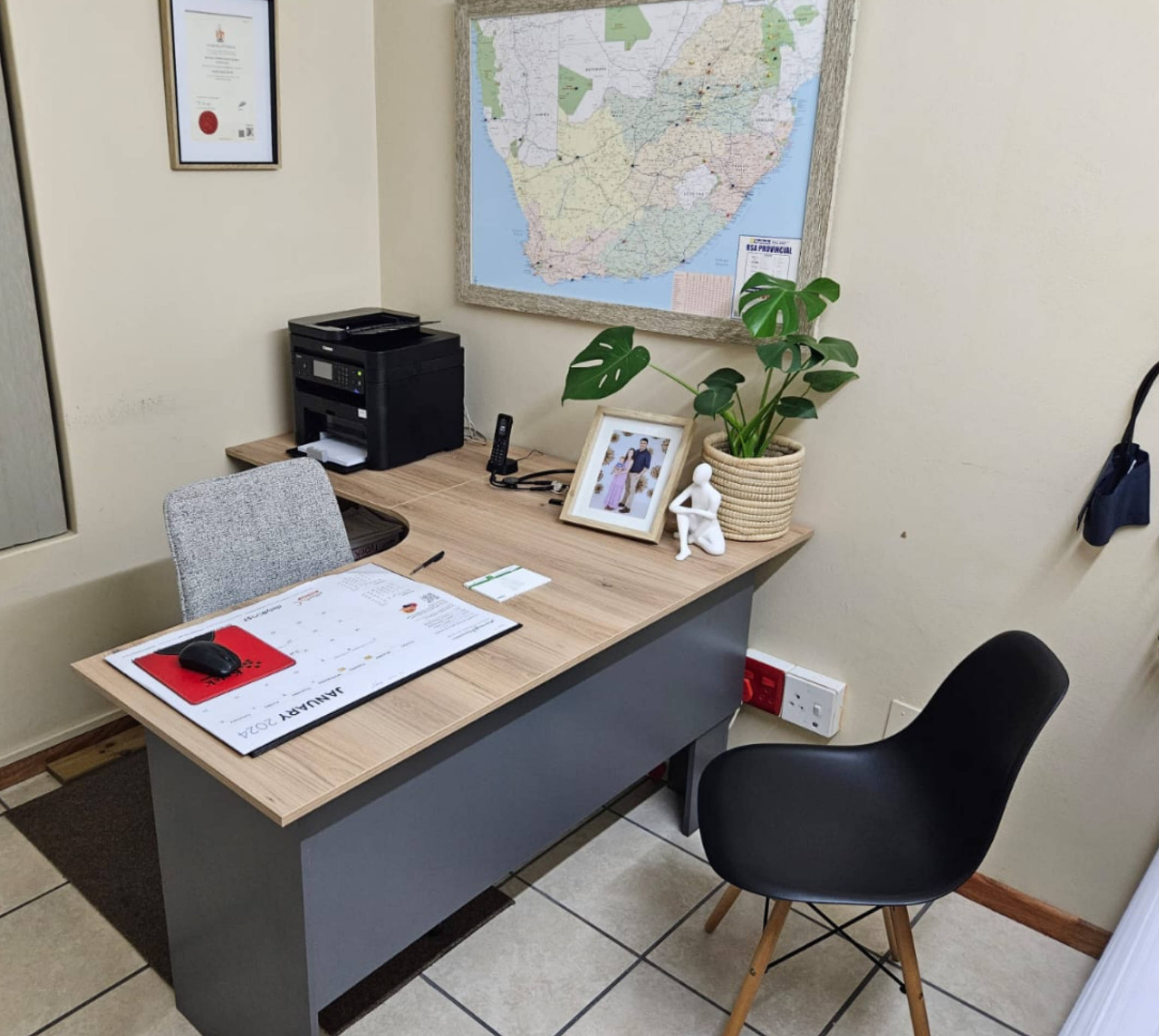 Small office desk with a calendar, mouse, printer, plant, framed photo, and two chairs on tiled floor.
