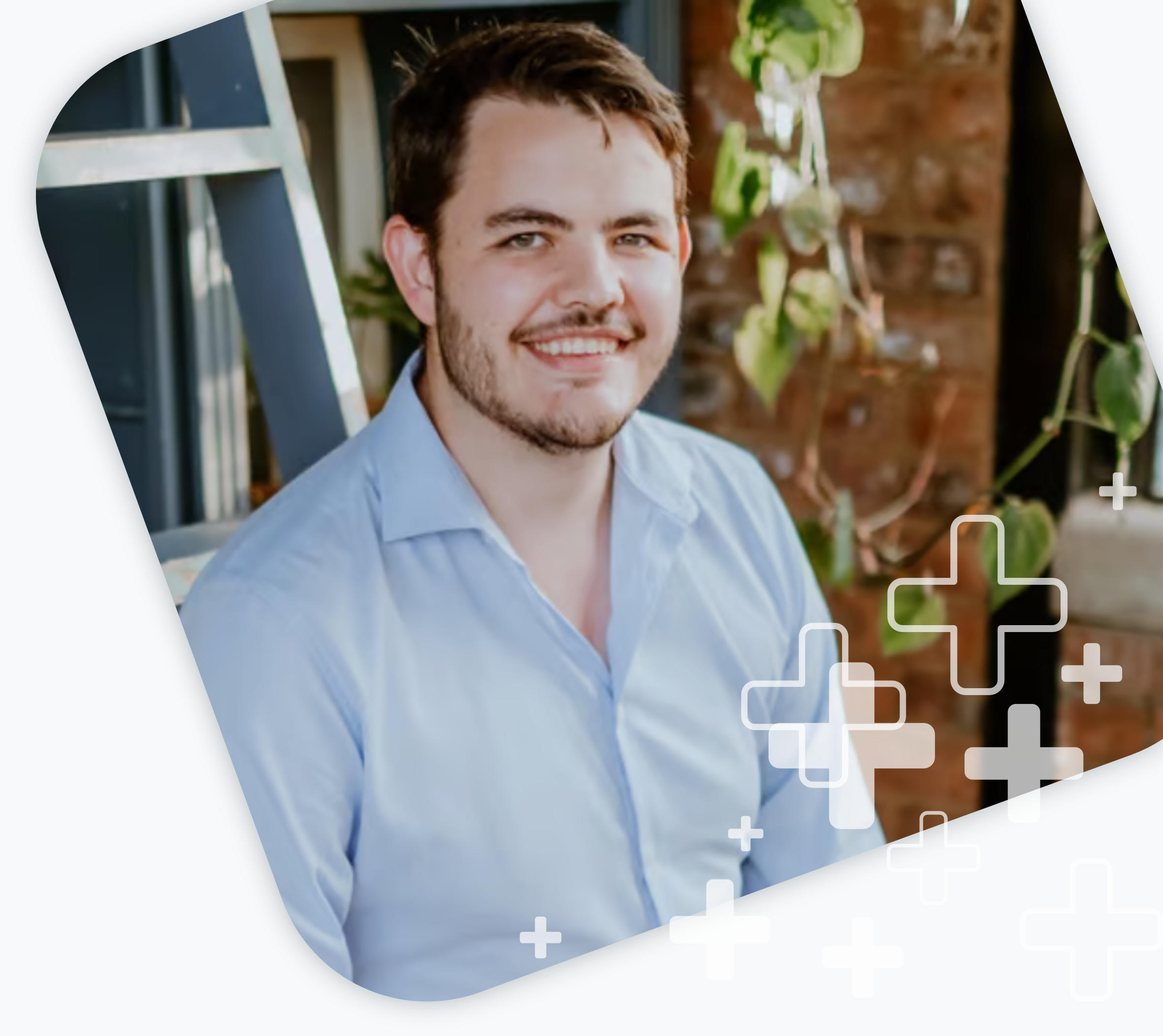 Smiling man in a light blue shirt sitting indoors with plants and a brick wall in the background.
