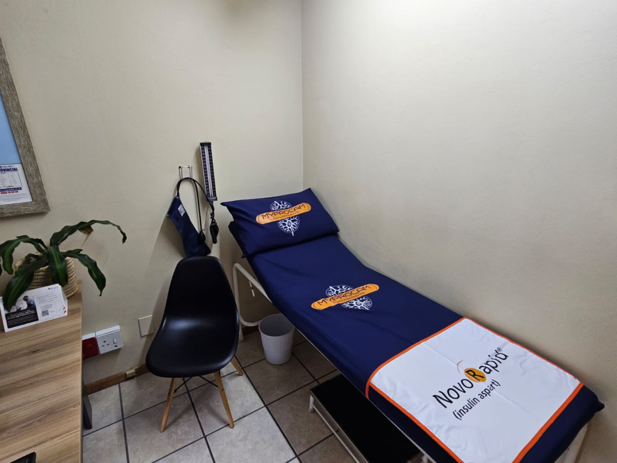 Medical examination room with a blue examination couch covered with NovaRapid (insulin aspart) branded fabric, a black chair, a stethoscope, blood pressure cuff on the wall, and a plant on a wooden cabinet.